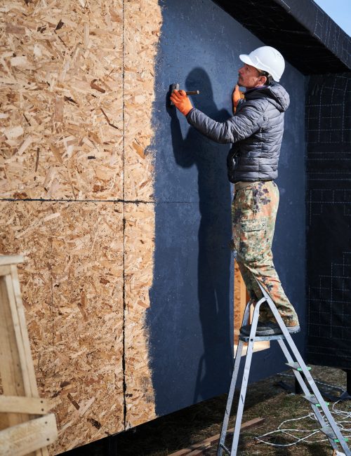 Male painter using paint roller, doing exterior paint work in a black color, standing on a ladder. Man worker building wooden frame house. Carpentry and construction concept.
